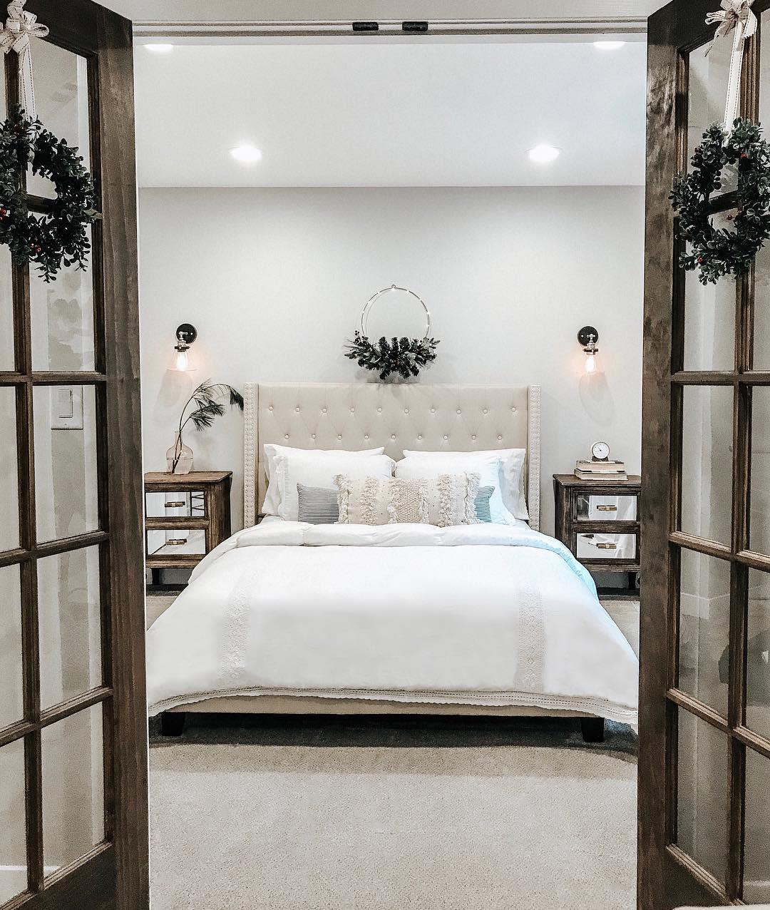 Cozy cottage-style bedroom with upholstered bed, rustic nightstands, and wreath decor.